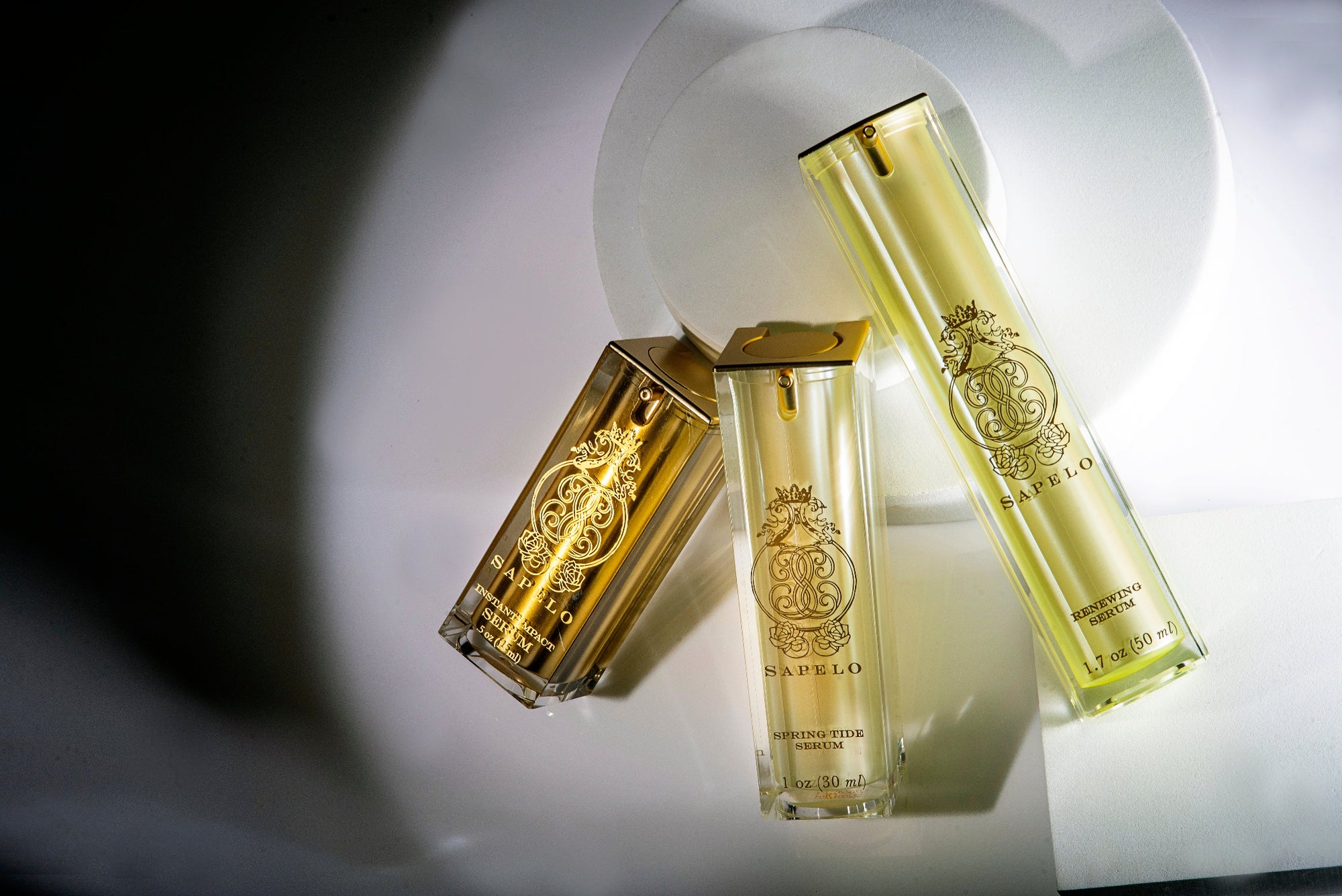 Three gold and clear skin care serum bottles on a reflective surface with a blurred background sapelo skin care 